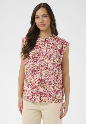 Kobieta ubrana w bezrękawnikową bluzkę na guziki w różowo-czerwony kwiatowy wzór oraz jasnobeżowe spodnie, stojąca na jednolitym tle.