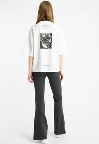 Kobieta z długimi włosami, ubrana w białą, oversizową koszulkę z abstrakcyjnym nadrukiem oraz czarne spodnie dzwony, stoi tyłem na białym tle.