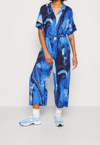 Mono azul mármol con mangas cortas, cuello y cinturón. Presenta un ajuste holgado y pantalones de longitud a la rodilla. Combinado con zapatillas de deporte azul claro.