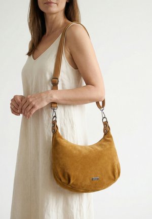 Femme en robe beige sans manches, portant un sac à bandoulière en daim jaune moutarde avec des sangles en cuir tanné ajustables.