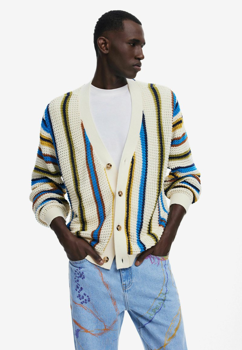 Cardigan lavorato a maglia di colore crema con strisce multicolori in blu, marrone e giallo. Presenta un collo a V, bottoni e polsini a costine. Abbinato a jeans blu chiaro.