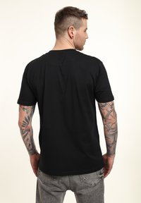 Černé tričko s krátkým rukávem, bavlněný materiál, kulatý výstřih, volný střih, jednoduchý design, na viditelné paži a rameni jsou vidět tetování.