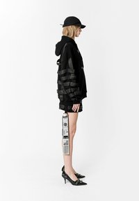 Sudadera negra de gran tamaño con detalles de cinta en las mangas, combinada con pantalones cortos negros y tacones puntiagudos. Los accesorios incluyen una gorra negra y una etiqueta colgante.