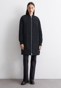 Veste bomber noire mi-longue avec fermeture éclair devant, poignets côtelés et poches latérales, fabriquée en tissu léger. Portée sur un pantalon et des chaussures sombres.