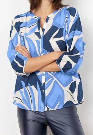Kobieta z założonymi rękami, ubrana w koszulę z abstrakcyjnym wzorem w odcieniach niebieskiego, białego i granatowego oraz ciemnoniebieskie skórzane spodnie.