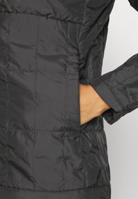 Main dans la poche d'une veste matelassée noire avec un motif carré visible et un poignet de manche.