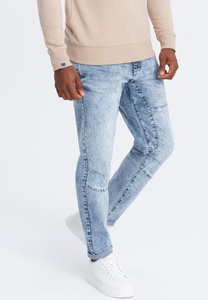 Jeans de mezclilla azul claro con un lavado desgastado, de corte tapered y detalles de patchwork. Combinados con zapatillas blancas y un sudadera beige.