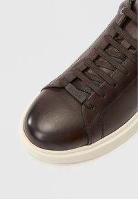 Baskets en cuir marron avec bout arrondi, lacets ton sur ton et étiquette logo sur le côté. Elles présentent une semelle en caoutchouc blanc et une texture lisse.