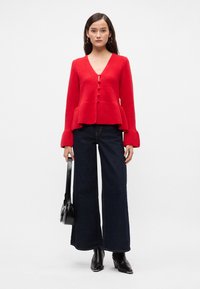 Cardigan tricotat roșu cu guler în V, închidere cu nasturi și tiv peplum. Purtați cu blugi negri evazați și botine cu toc negre.