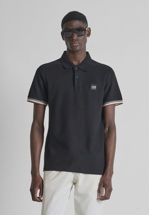 Uomo con polo nera con toppa con logo e pantaloni bianchi, in piedi contro uno sfondo grigio uniforme, indossa grandi occhiali da sole neri.