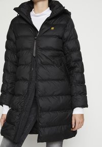 Nainen, joka on pukeutunut pitkään mustaan tikattuun talvitakkeeseen, jossa on huppu, vetoketjukiinnitys ja sivutaskut, valkoisen poolopaidan ja harmaiden farkkujen päällä.