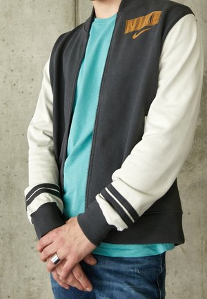 Personne portant une veste Nike noire et blanche par-dessus un t-shirt turquoise, un jean bleu et une bague en argent à la main gauche devant un mur gris.