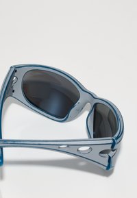 Zilas un pelēkas saulesbrilles ar tumšiem stikliem, kas izceļas ar elegantu, sportisku dizainu un strukturētām kājām ar vairākiem izgriezumiem.