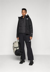 Zwarte geïsoleerde jas over een puffer zwarte vest, gecombineerd met donkere cargobroeken. Draagt een lichtgrijze rugzak met zwarte accenten.