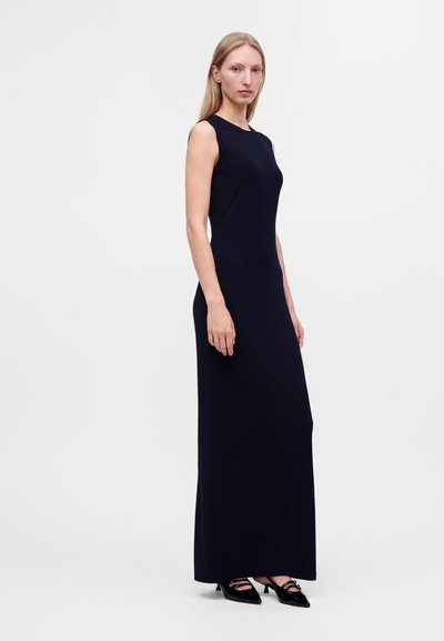 Femme debout portant une robe noire sans manches, longue jusqu'au sol, et des talons noirs à bout pointu sur un fond blanc uni.