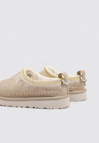 Chaussures slip-on en suede beige avec des coutures jaunes et blanches, doublure en laine, semelles robustes beige et languettes à l'arrière.