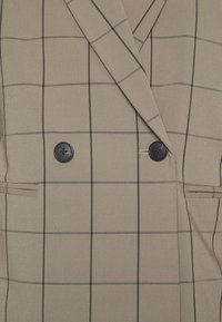 Blazer beige avec un motif discret à carreaux fenêtre, double boutons noirs et col à revers cranté.
