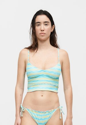 Γυναίκα που στέκεται με το πρόσωπο προς τα εμπρός, φοράει μπλε και κίτρινο ριγέ τοπ μπικίνι και αντίστοιχα μπικίνι με κορδόνια στο πλάι σε λευκό φόντο.