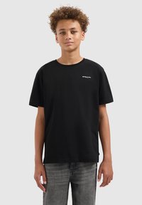 Tienerjongen met krullend haar, gekleed in een zwart 'Ballin'' t-shirt en grijze jeans, staat tegen een effen lichtgrijze achtergrond.