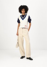 White cable-knit vest with blue accents over navy long-sleeve top, paired with light beige wide-leg pants and black shoes. Red embroidery detail.