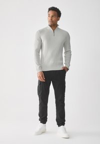 Suéter de canalé gris claro con diseño de media cremallera, combinado con pantalones cargo negros que tienen bolsillos laterales y zapatillas blancas. Fondo simple.