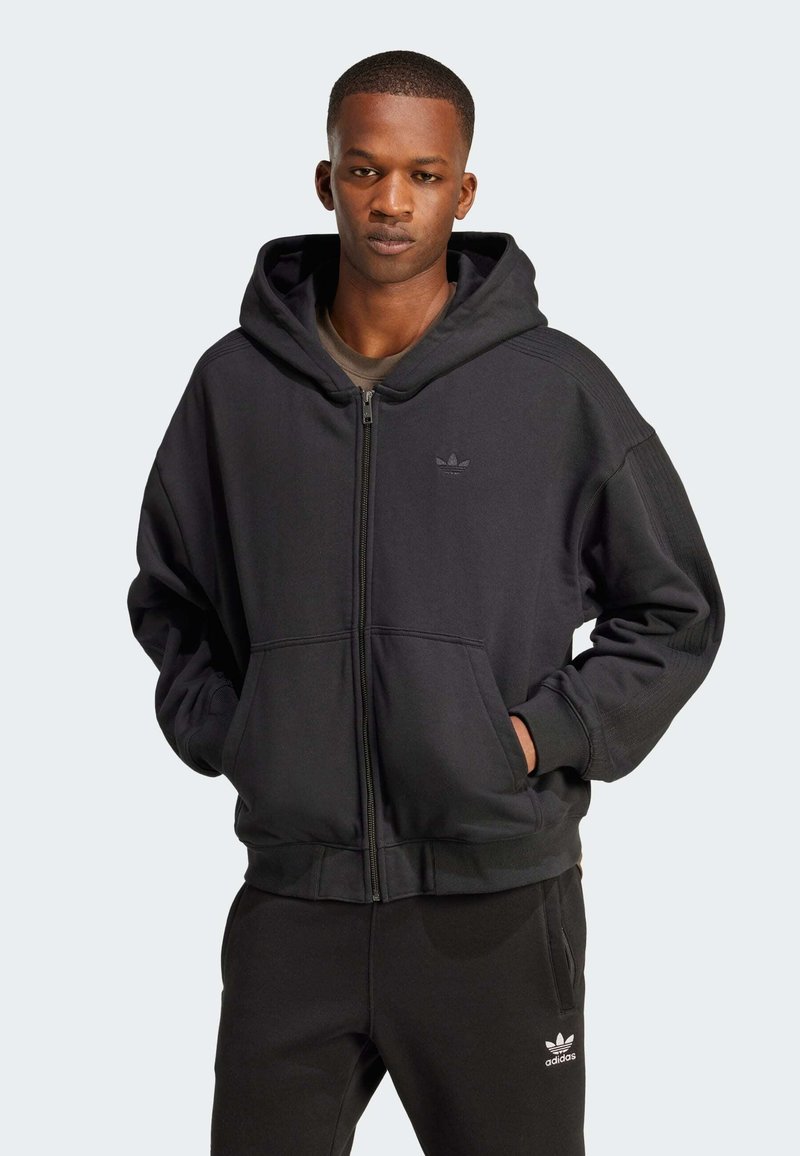 Melnā kapučja jaka ar rāvējslēdzēju, izgatavota no flīsa, ar lielu kapuci, priekšējām kabatām un Adidas logo uz krūtīm un biksēm. Gluda tekstūra un brīvs piegriezums.