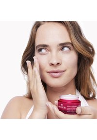NUXE Merveillance crème in een kleine, doorzichtige pot met een heldere roze basis en een witte deksel. Een hand brengt crème aan op het gezicht, waarbij een gladde textuur wordt weergegeven.