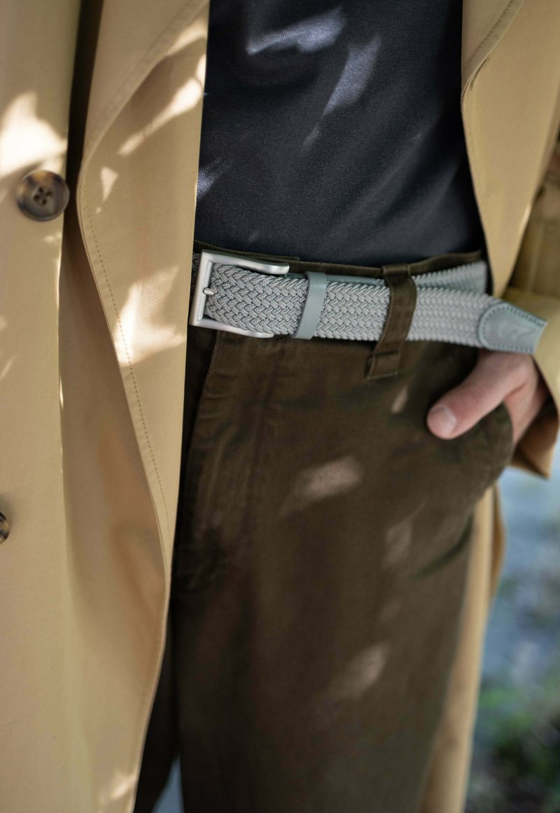 Beige Trenchcoat über einem dunklen Hemd, kombiniert mit dunkelgrünen Hosen. Ein hellgraßer, gewebter Gürtel mit einer silbernen Schnalle und Lederakzenten.