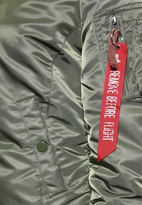 Olivgrön nylonjacka med quiltad textur, prydd med en röd tygetikett som står "REMOVE BEFORE FLIGHT" och en svart knapppdetail.