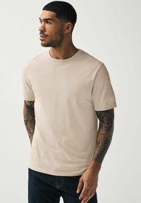 Camiseta de cuello redondo beige de tejido de algodón, de manga corta, con un corte relajado. Tatuaje del tono de piel visible en el brazo izquierdo.