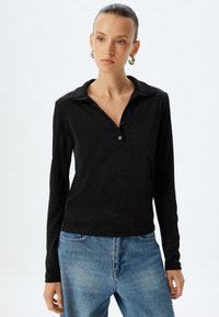Schwarzes Langarm-Polohemd mit weicher Textur, Knopfverschluss und klassischem Kragen, kombiniert mit hellblauen Jeans.