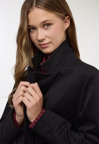 Cappotto in pile nero con collo alto, vestibilità aderente e tessuto dalla trama lavorata. Sotto, si intravede il polsino di una camicia a righe rosse, che aggiunge un dettaglio.