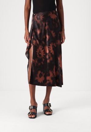 Femme portant un haut noir avec une jupe midi fendue à fleurs noire et marron et des sandales noires à talons blocs ouvertes à la boucle, debout devant un fond blanc.