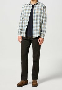 Hombre vestido con zapatos de ante marrón, jeans negros, camiseta azul marino y camisa de cuadros beige-azul, de pie contra un fondo liso.
