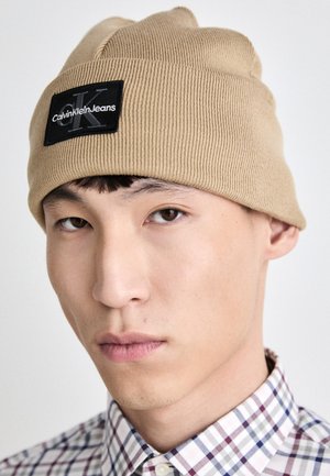 Bež pletena kapa z črno nalepko, na kateri je logotip "Calvin Klein Jeans". Model v karirasti srajci, z bližnjim posnetkom na klobuku.