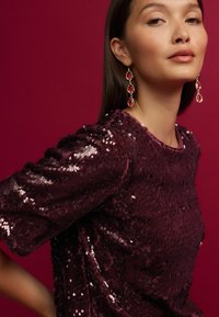 Top en sequins bourgogne avec des manches courtes bouffantes et un col rond, accompagné de boucles d'oreilles pendantes dorées et rouges. Fond en rouge foncé uni.