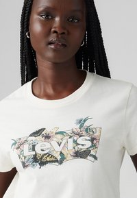 Tricou alb cu mânecă scurtă, având un grafic floral colorat cu fluturi și logo-ul "Levi's" în centru. Textură netedă din bumbac.