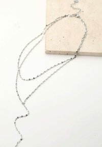 Collier en argent avec plusieurs chaînes reliées, de longueurs et d'épaisseurs variées, reposant sur une surface en pierre de couleur claire.