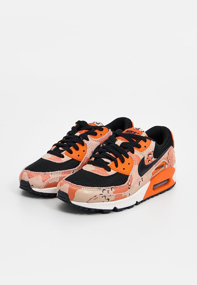 Zapatillas Nike con una mezcla de patrones de camuflaje en naranja, negro y beige. Sus características incluyen una parte superior de malla negra y una suela blanca con amortiguación de aire.