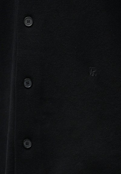 Μαύρο πλεκτό ύφασμα με τρία μαύρα κουμπιά σε κατακόρυφη διάταξη και διακριτικό κεντητό γράμμα "R" στη δεξιά πλευρά.