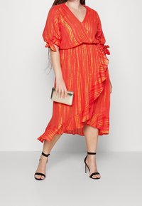 Rotes Kleid mit goldenen vertikalen Streifen, V-Ausschnitt, kurzen Ärmeln mit Schleifen, volantsaum, kombiniert mit schwarzen Riemchenschuhen und einer metallischen Clutch.