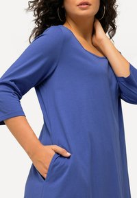 Blauwe katoenen jurk met een ronde halslijn, driekwart mouwen en zijzakken. Soepele textuur en een ontspannen pasvorm met minimale details.