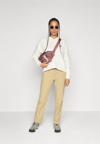 Kvinne med lange fletter iført solbriller, hvit hettegenser, beige bukser, tursko og en mauve crossbody-veske, stående mot en ensfarget bakgrunn.