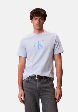 Joven con cabello rizado castaño que lleva una camiseta azul claro con el logo de Calvin Klein y jeans negros, de pie contra un fondo liso.