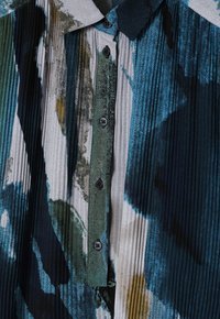 Blumenbedruckte plissierte Bluse mit einer Mischung aus Blau-, Grün- und Gelbtönen, mit einem Kragen und schwarzen Knöpfen entlang der Vorderseite.