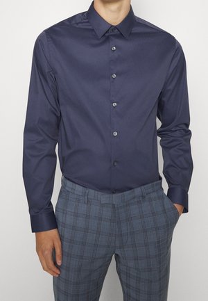 Mężczyzna noszący dopasowaną ciemnoniebieską koszulę na guziki, włożoną do szarych kraciastych spodni, z jedną ręką w kieszeni.