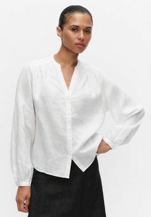 Kvinde med opsat hår iført en hvid skjorte med knapper og sort nederdel, stående med den ene hånd på hoften, neutral baggrund.