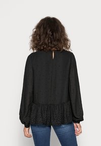 Femme avec des cheveux bouclés portant un blouse à manches longues noire à motif à pois et avec un ourlet à volants, associée à un jean bleu, vue de dos.