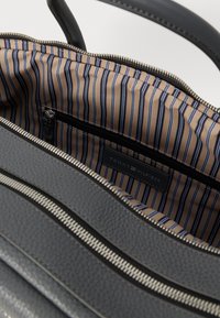 Sac en cuir texturé gris avec des zippers argentés et une doublure rayée beige. Comprend une poche zippée et une étiquette avec le logo "TOMMY HILFIGER".