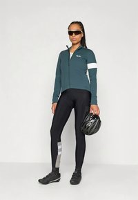 Turkooise fietsjacks met witte accenten, zwarte fietsbroek met grijze strepen, en zwarte helm in de rechterhand. Gestructureerde stoffen.